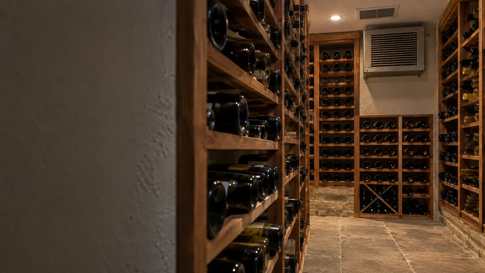A wine cellar with a ceiling supply grille and white plaster walls, seen close and low along the racks.