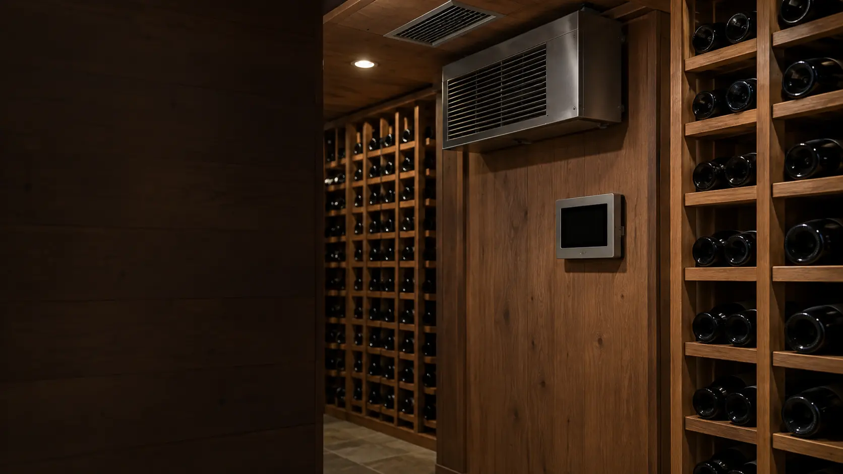 A wall-mounted digital controller for a cellar cooling unit on a wood-paneled wine cellar wall, its small screen completely dark, a ceiling supply grille above and rows of unlabeled bottles in wooden racks to the side.