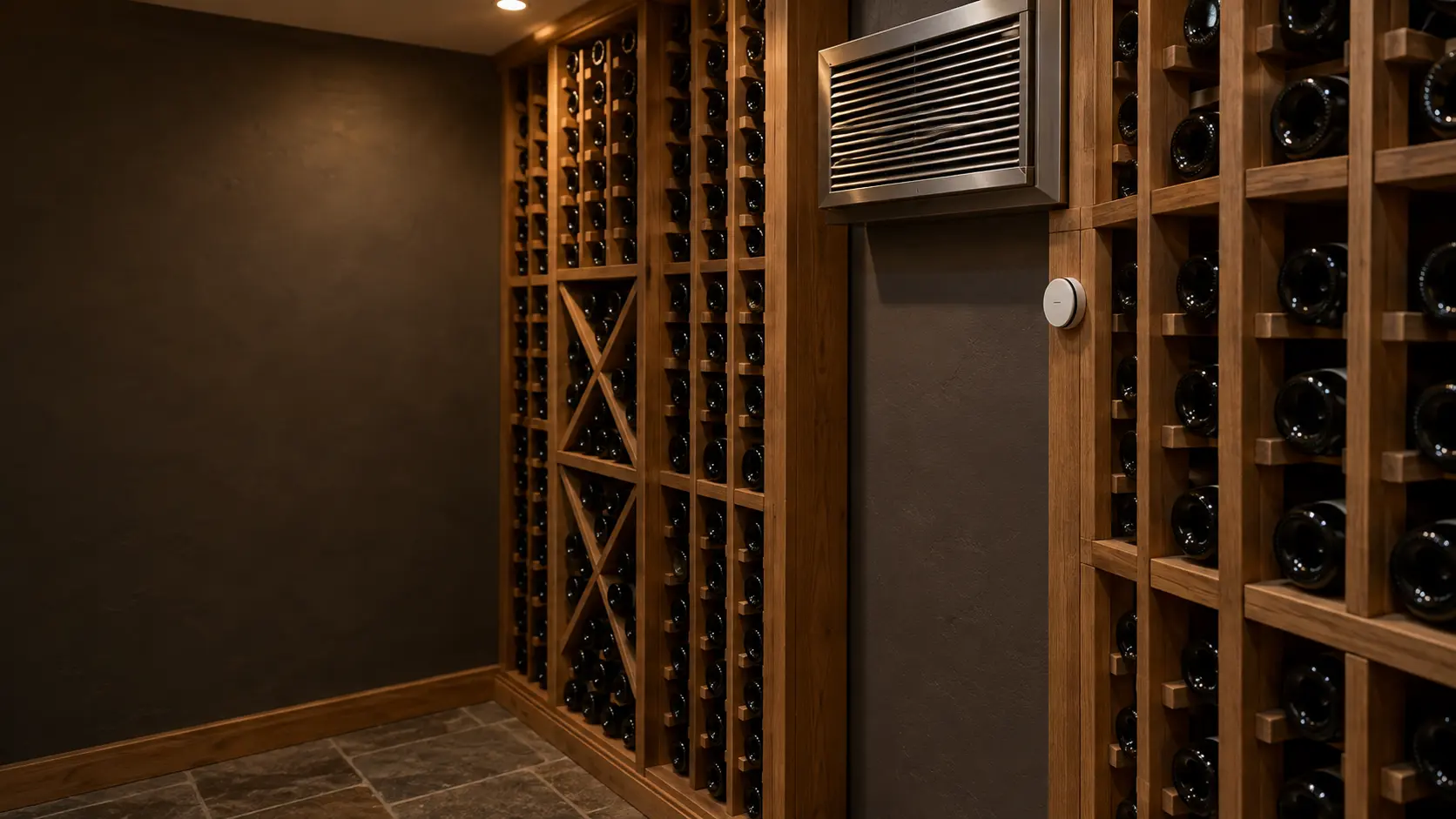 A compact wine cellar with a through-the-wall cooling unit grille high on a painted wall and a small round temperature sensor mounted on a rack post just below it, unlabeled bottles lying in wooden racks.
