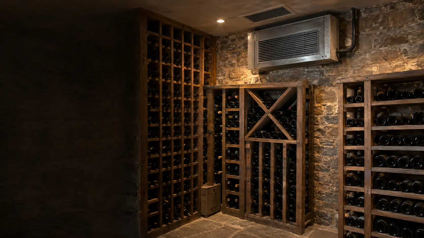The outdoor condenser of a split wine cellar cooling system beside a house wall, its insulated line set running up into the siding, the fan guard on top and the fins dulled by weather.