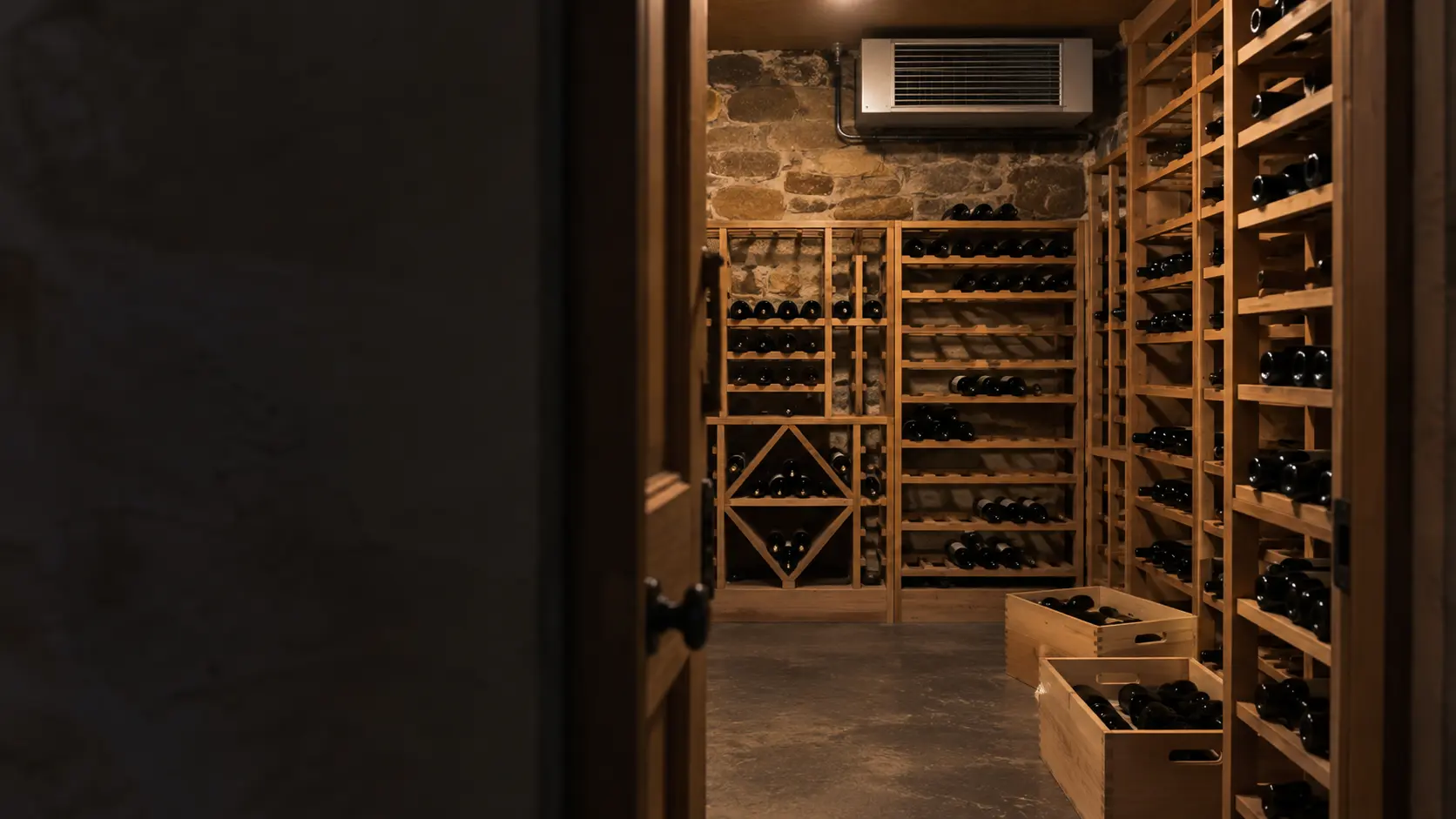The doorway of a stone-walled wine cellar with a split-system evaporator mounted high on the back wall, a few open wooden cases on the floor holding bottles lying on their sides, racks behind them half full.